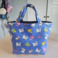Load image into Gallery viewer, A blue fabric tote bag with a repeating pattern of colorful open books, shown hanging against a beige wall in an indoor setting.
