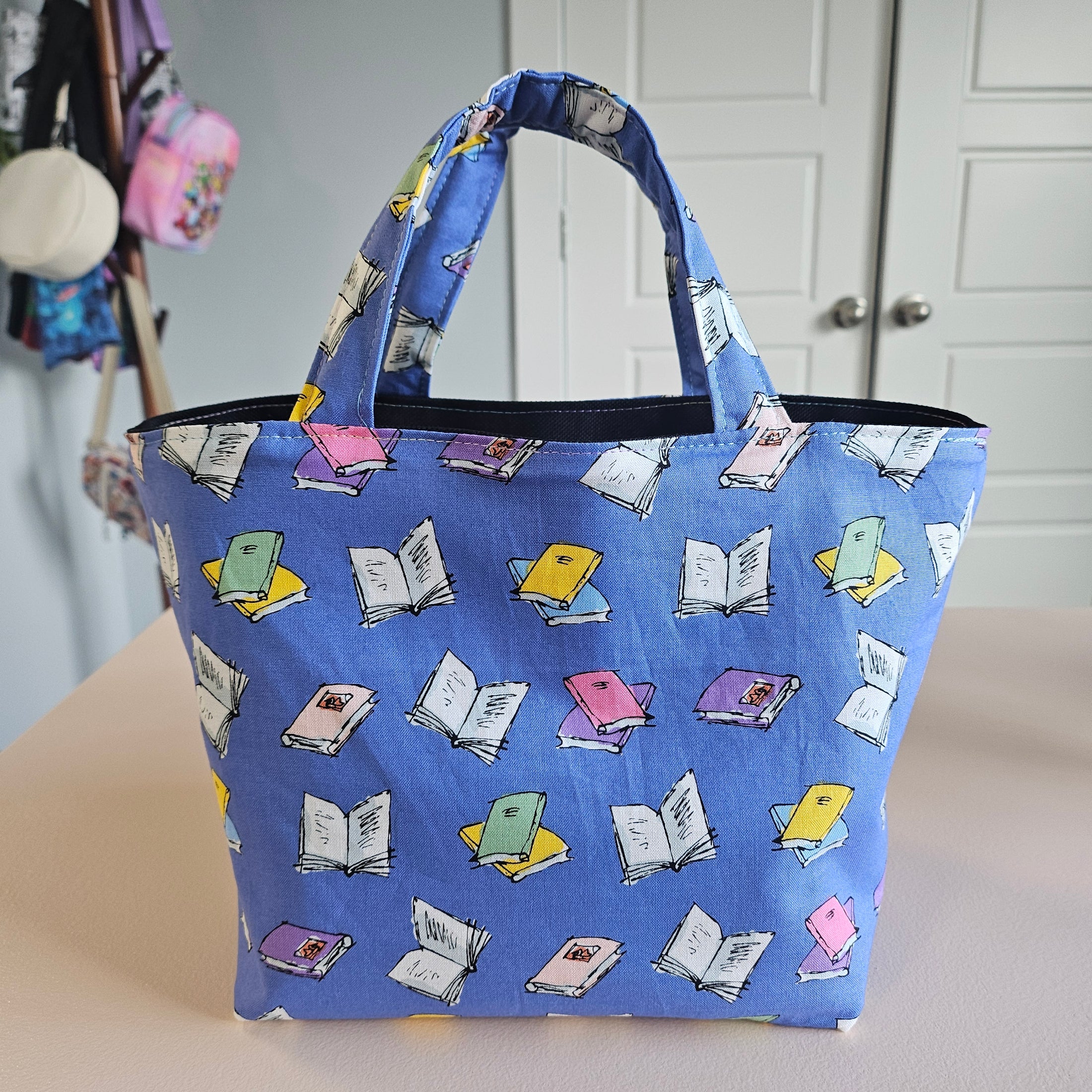 A blue fabric tote bag with a repeating pattern of colorful open books, shown hanging against a beige wall in an indoor setting.