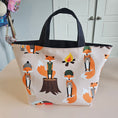 Load image into Gallery viewer, A fabric tote bag with a cream background and an all-over print of orange foxes in different activities, featuring black canvas handles, shown hanging against a wall.
