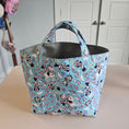 Load image into Gallery viewer, A light blue fabric tote bag with a repeating cartoon Minnie Mouse pattern, hanging on a beige wall next to white cabinets.
