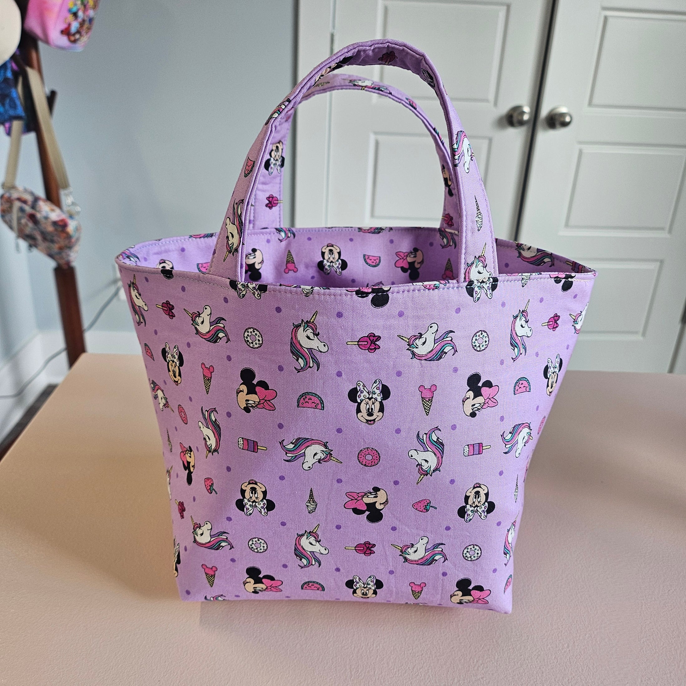 A light purple fabric tote bag with a repeating cartoon Minnie Mouse and Mickey Mouse pattern, hanging against a beige wall next to white paneled cabinets.