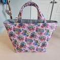 Load image into Gallery viewer, A pink fabric tote bag with a colorful floral and basketweave pattern, featuring gray handles and lining, photographed hanging against a beige wall with white cabinets in the background.
