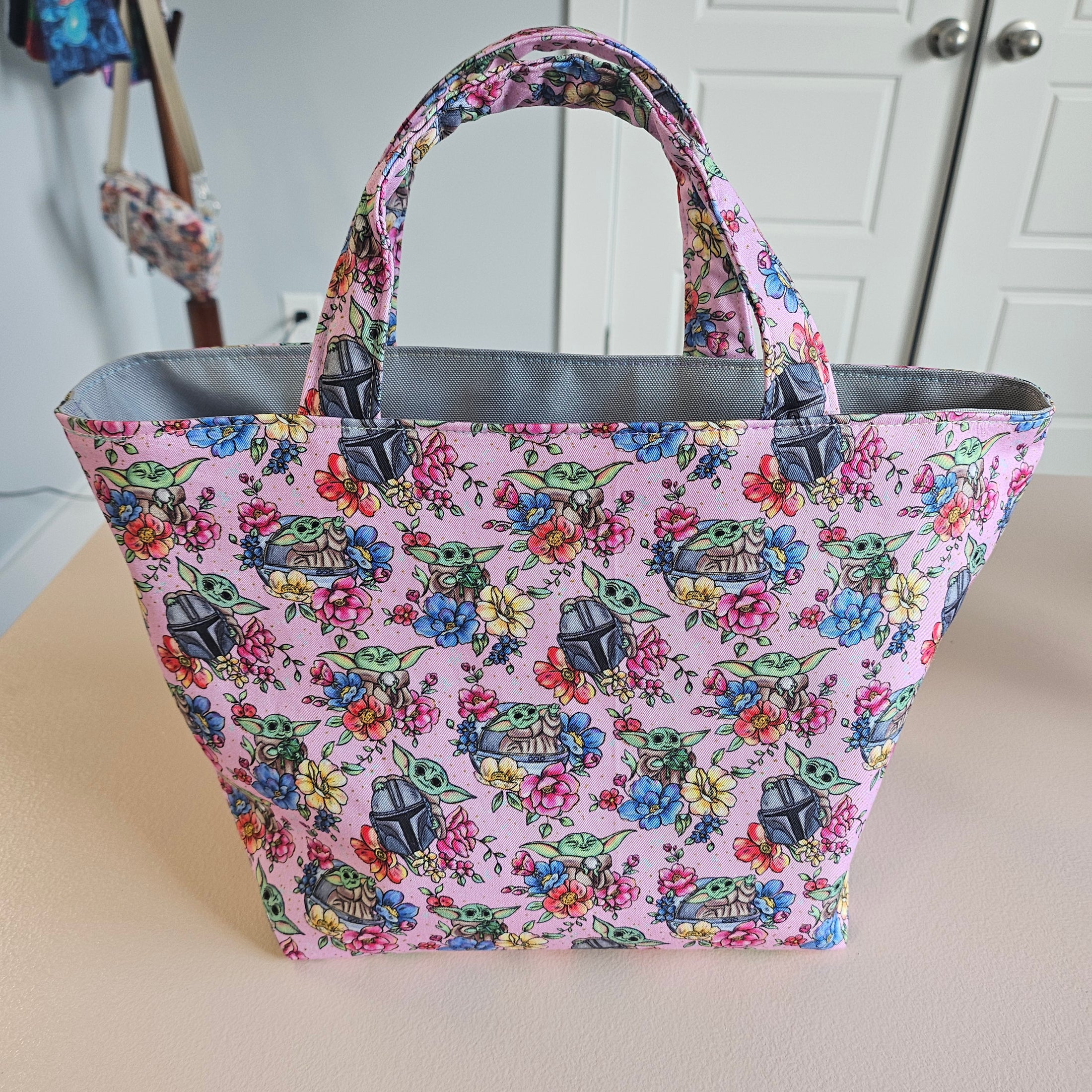 A pink fabric tote bag with a colorful floral and basketweave pattern, featuring gray handles and lining, photographed hanging against a beige wall with white cabinets in the background.