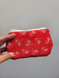 Load image into Gallery viewer, A hand holding a small zippered pouch made of red fabric with a white repeating pattern of sewing needles and thread, against a plain gray background.
