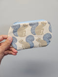 Load image into Gallery viewer, A hand holding a small zippered pouch with a repeating pattern of beige squirrels and blue-gray tails on a light background, against a plain gray backdrop.
