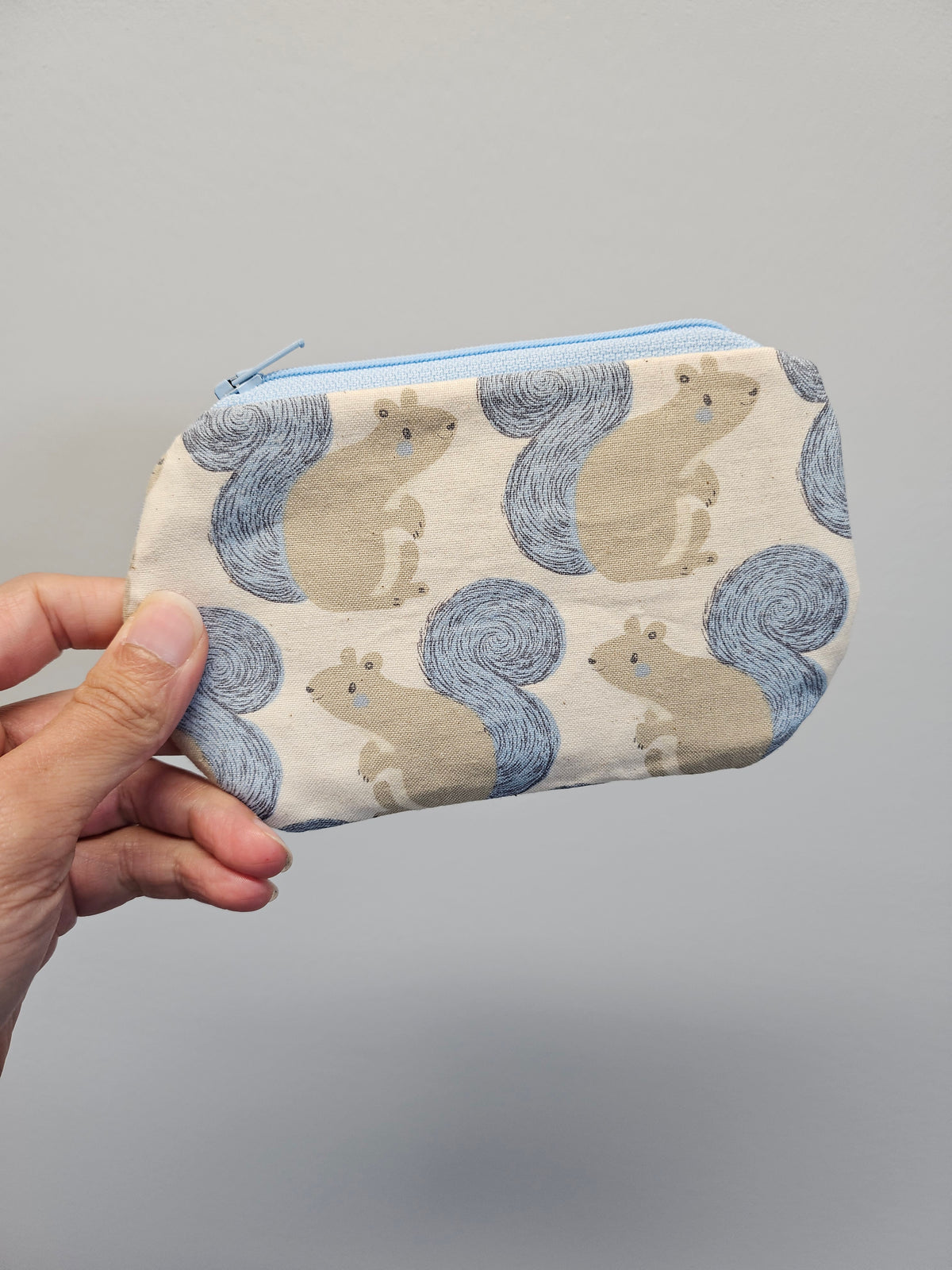 A hand holding a small zippered pouch with a repeating pattern of beige squirrels and blue-gray tails on a light background, against a plain gray backdrop.