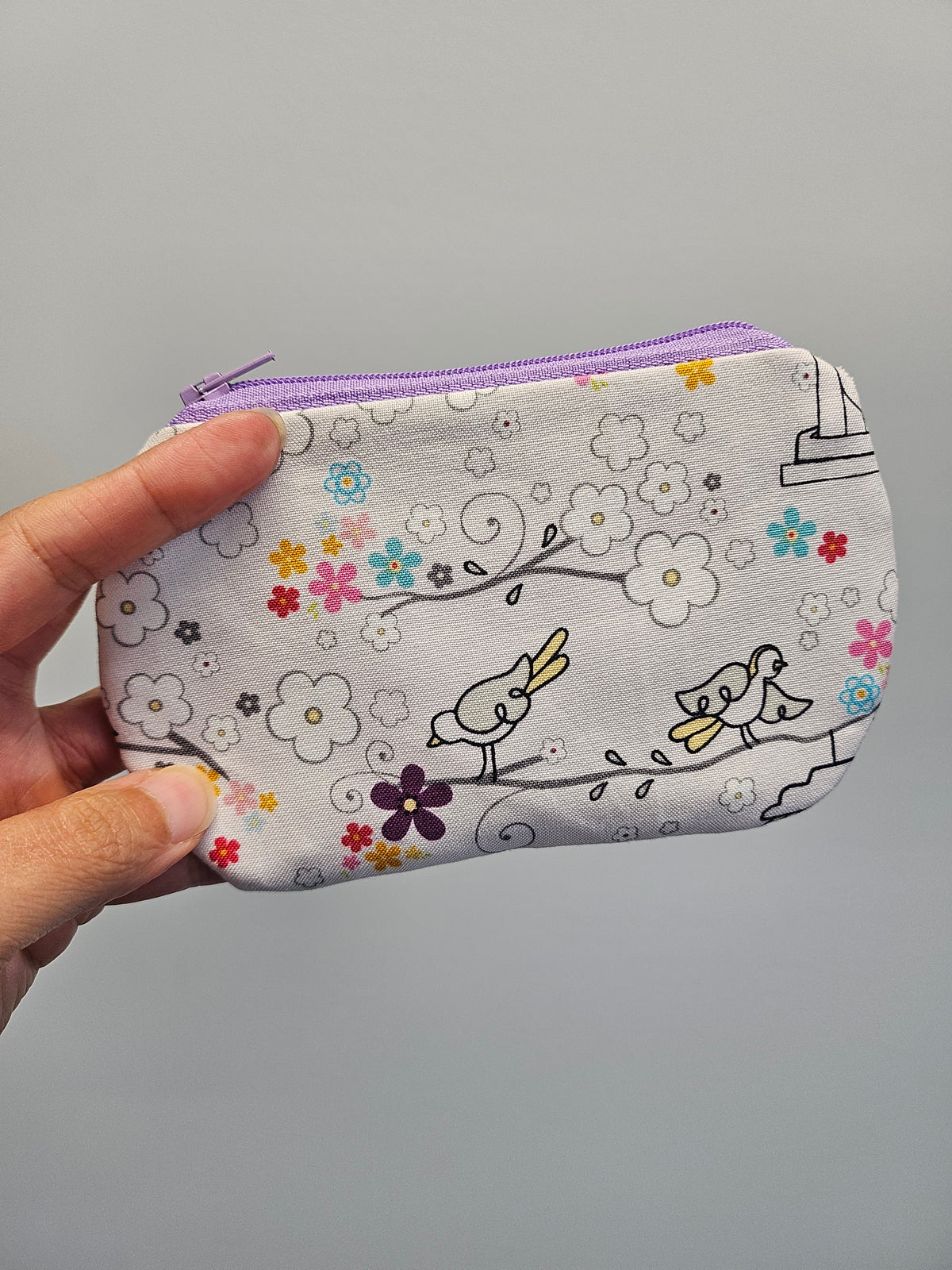 A hand holding a small zippered pouch with a light gray background and a whimsical print of birds, flowers, and branches. The pouch has a lavender purple zipper.