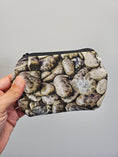 Load image into Gallery viewer, A hand holding a small zippered pouch with an all-over print of smooth river stones in earthy gray, beige, brown, and white tones, against a plain light gray background.

