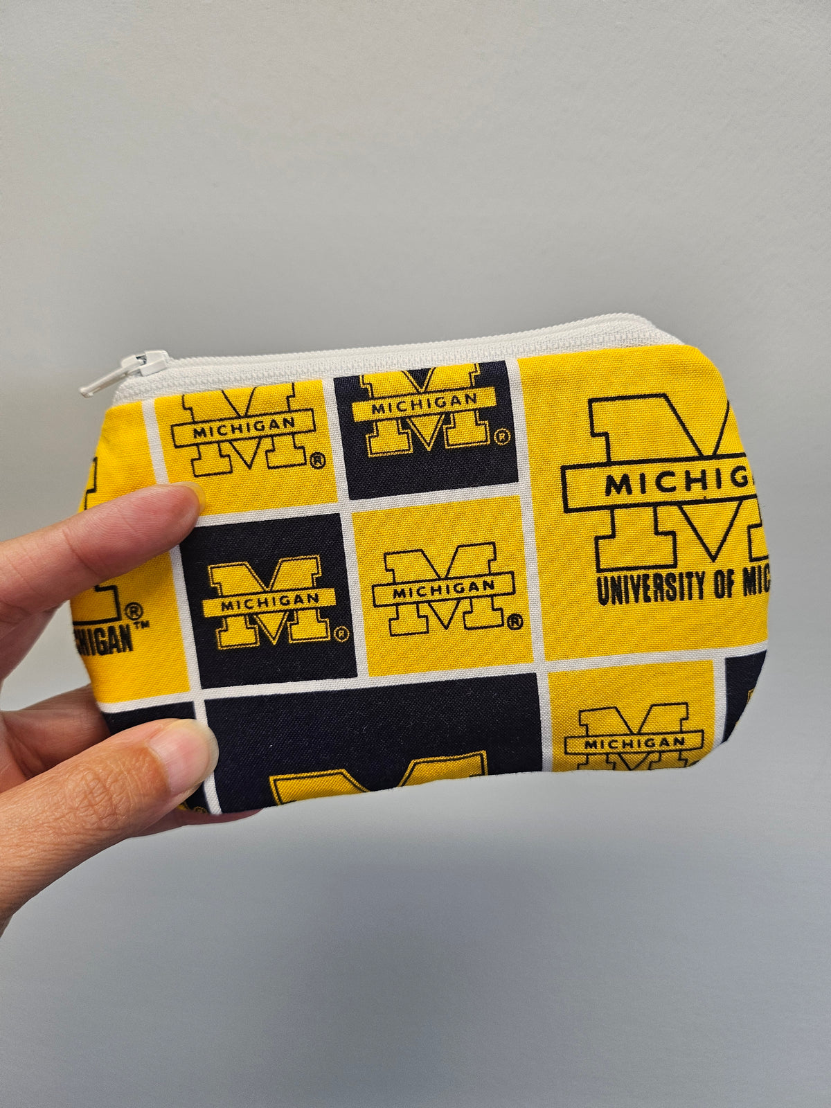 Hand holding a university of michigan mini zipper pouch against a light blue wall.