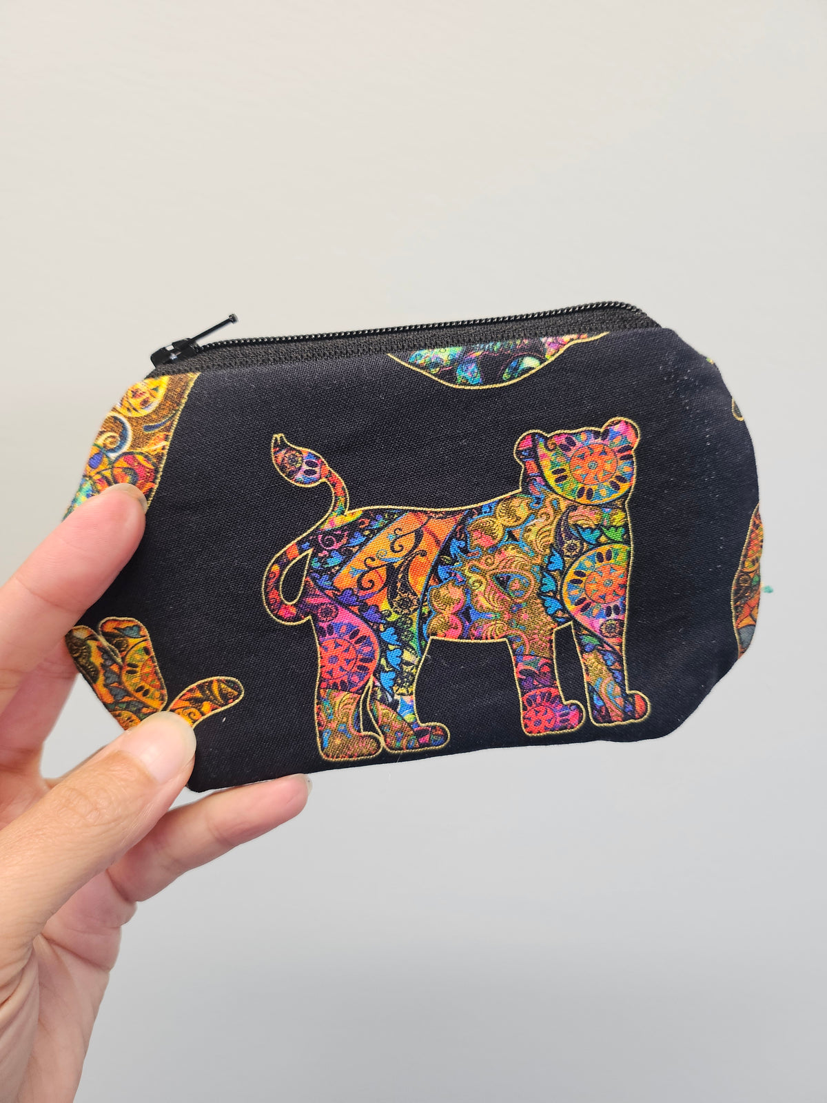 A hand holding a small zippered pouch with a black background and a colorful, patterned jaguar design. The pouch features a black base with a vibrant, multicolored jaguar motif outlined in gold, set against a plain light gray background.