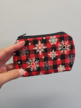 Load image into Gallery viewer, A hand holding a small zippered pouch with a red and black buffalo plaid pattern decorated with white and silver snowflakes, set against a plain light gray background.
