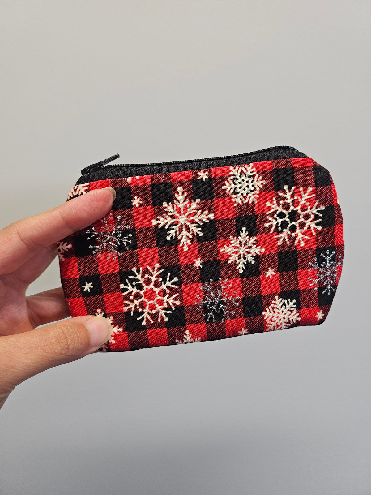A hand holding a small zippered pouch with a red and black buffalo plaid pattern decorated with white and silver snowflakes, set against a plain light gray background.