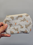 Load image into Gallery viewer, A hand holding a small zippered fabric pouch with a print of gray bunnies eating orange carrots on a light gray background.
