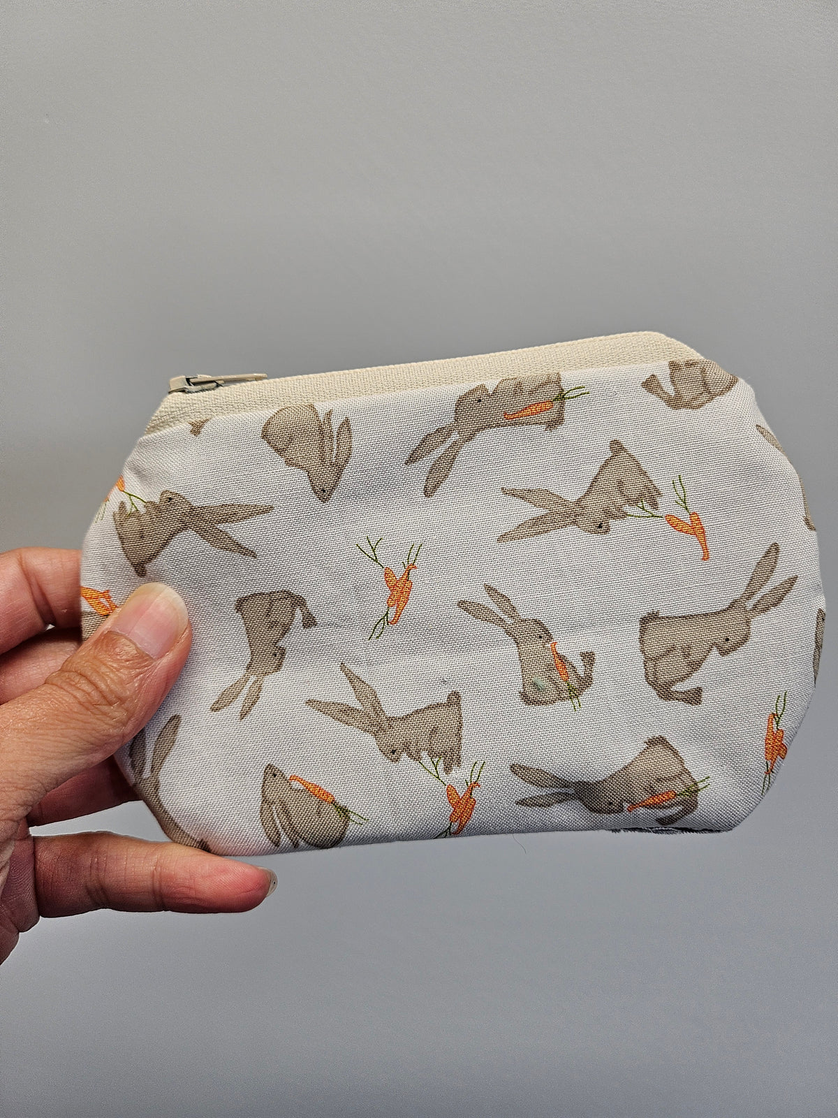 A hand holding a small zippered fabric pouch with a print of gray bunnies eating orange carrots on a light gray background.