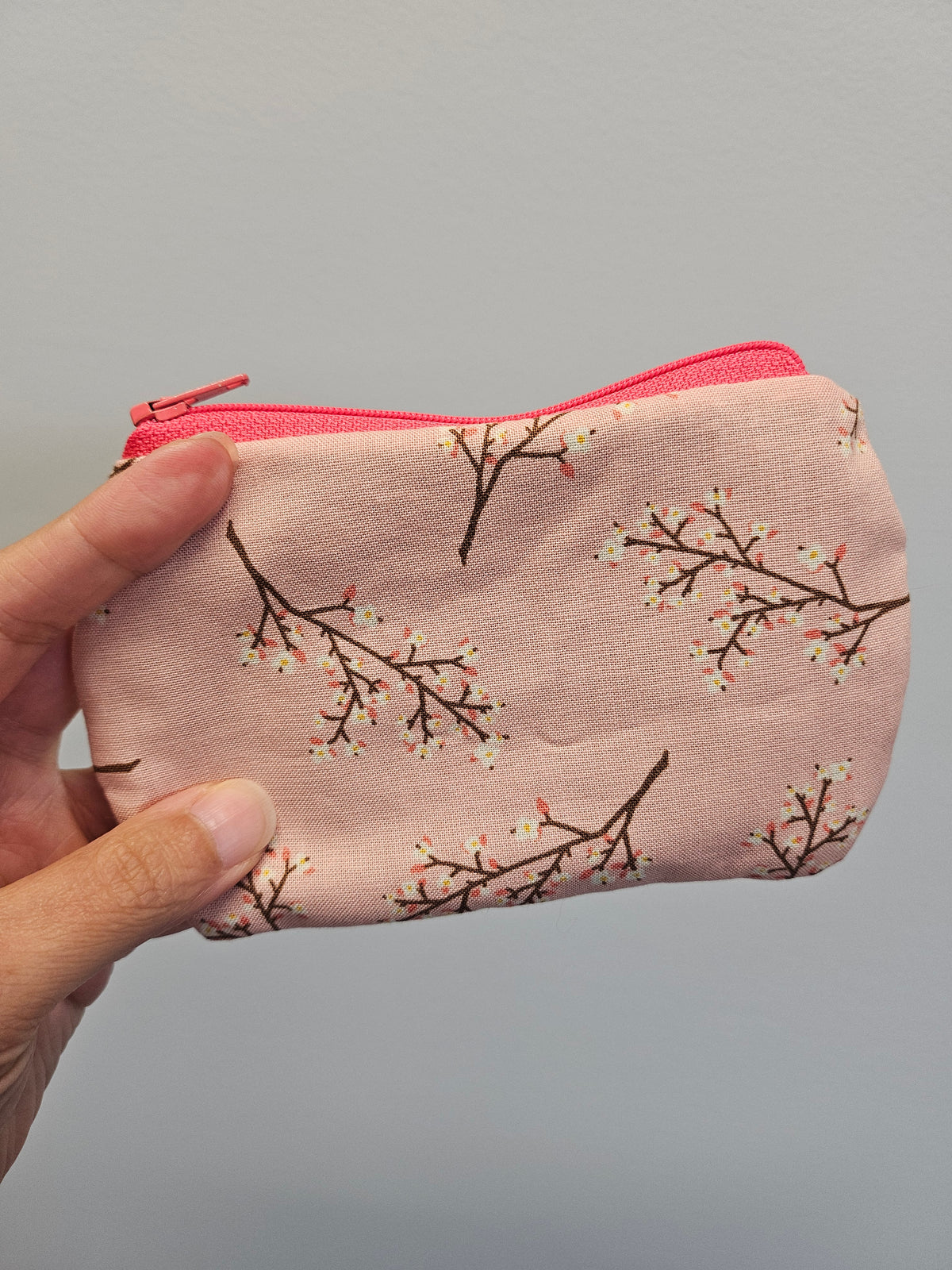 A hand holding a small zippered pouch with a pink background and a cherry blossom branch pattern, featuring a bright pink zipper.