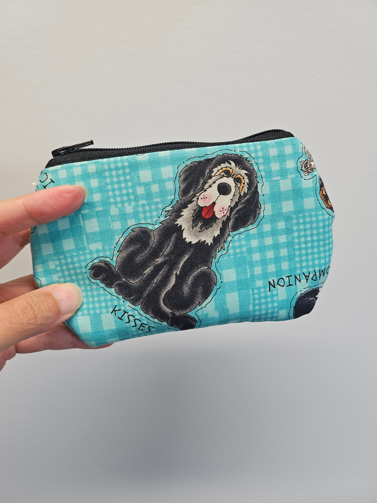 Hand holding a turquoise zipper pouch against a gray wall. The zipper pouch has images of dogs and cats on it.
