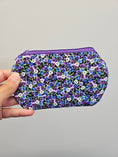 Load image into Gallery viewer, Hand holding a small zipper pouch against a light gray wall. The zipper pouch is purple, blue and white with pansies on it.
