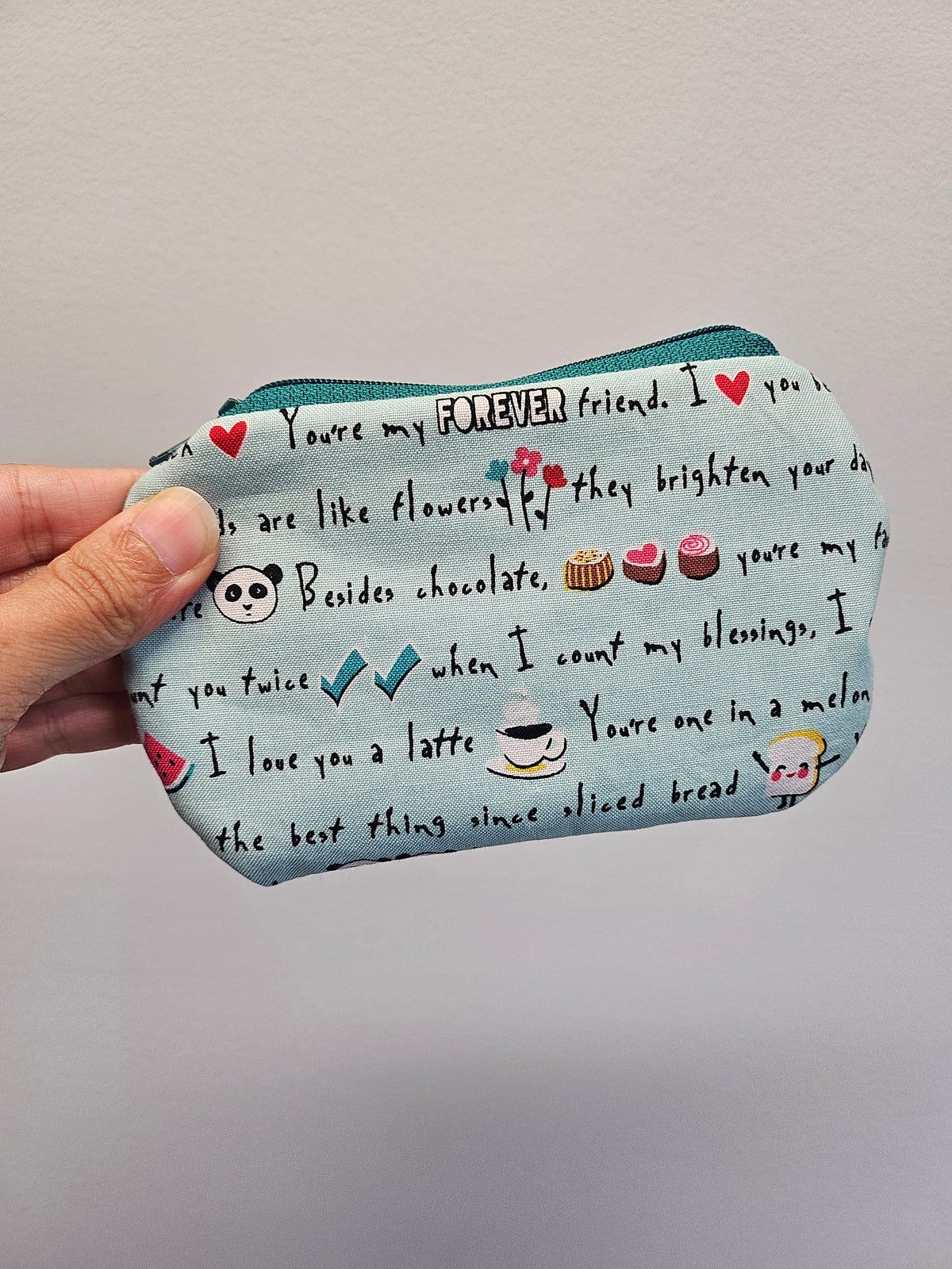 A hand holding a small zippered pouch with a light blue background printed with black text, red hearts, and small cute illustrations like a panda face, flowers, cupcakes, and a coffee cup, against a plain light gray wall.
