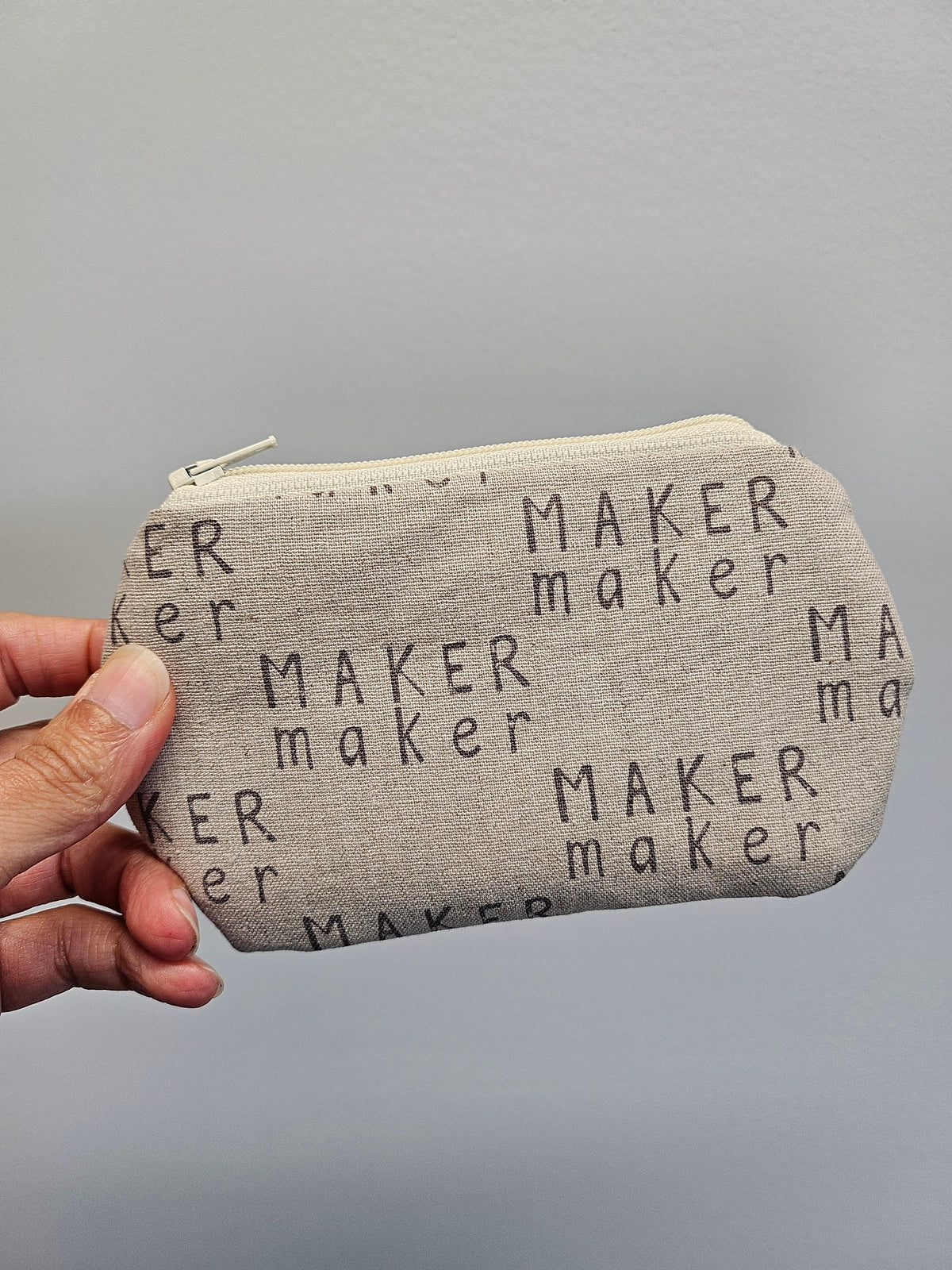 A hand holding a small zippered pouch made of light beige fabric printed with the word 'MAKER maker' in black text, against a plain gray background.