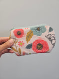 Load image into Gallery viewer, A hand holding a small zippered pouch with a floral print. The pouch features pink and red flowers, green and blue leaves, and black script text reading 'Adios Bitchachos' on a light gray background.
