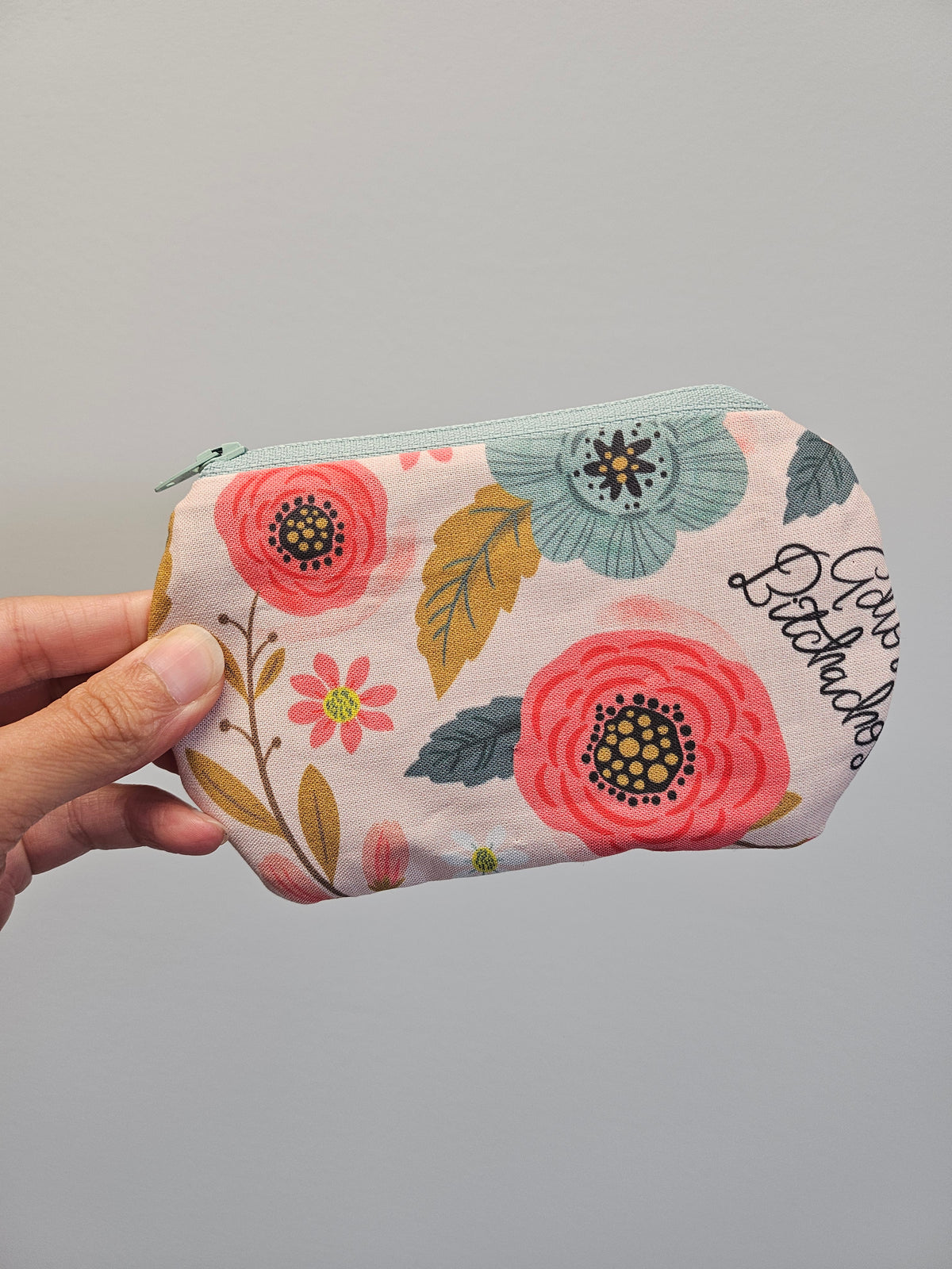 A hand holding a small zippered pouch with a floral print. The pouch features pink and red flowers, green and blue leaves, and black script text reading 'Adios Bitchachos' on a light gray background.