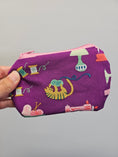 Load image into Gallery viewer, A hand holding a small zippered pouch with a purple background and a colorful sewing-themed print featuring a yellow cat, spools of thread, yarn balls, and sewing machines.
