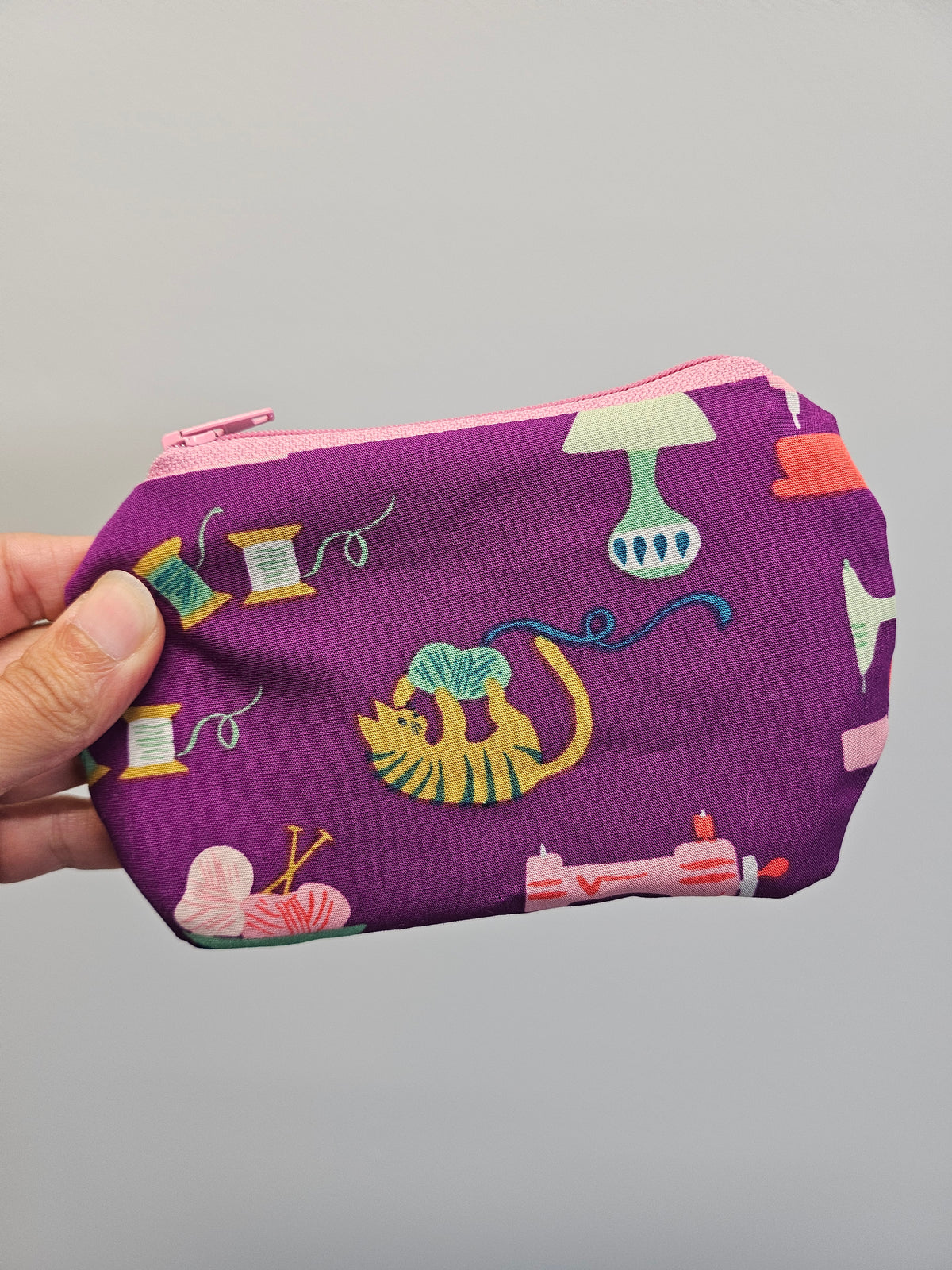 A hand holding a small zippered pouch with a purple background and a colorful sewing-themed print featuring a yellow cat, spools of thread, yarn balls, and sewing machines.