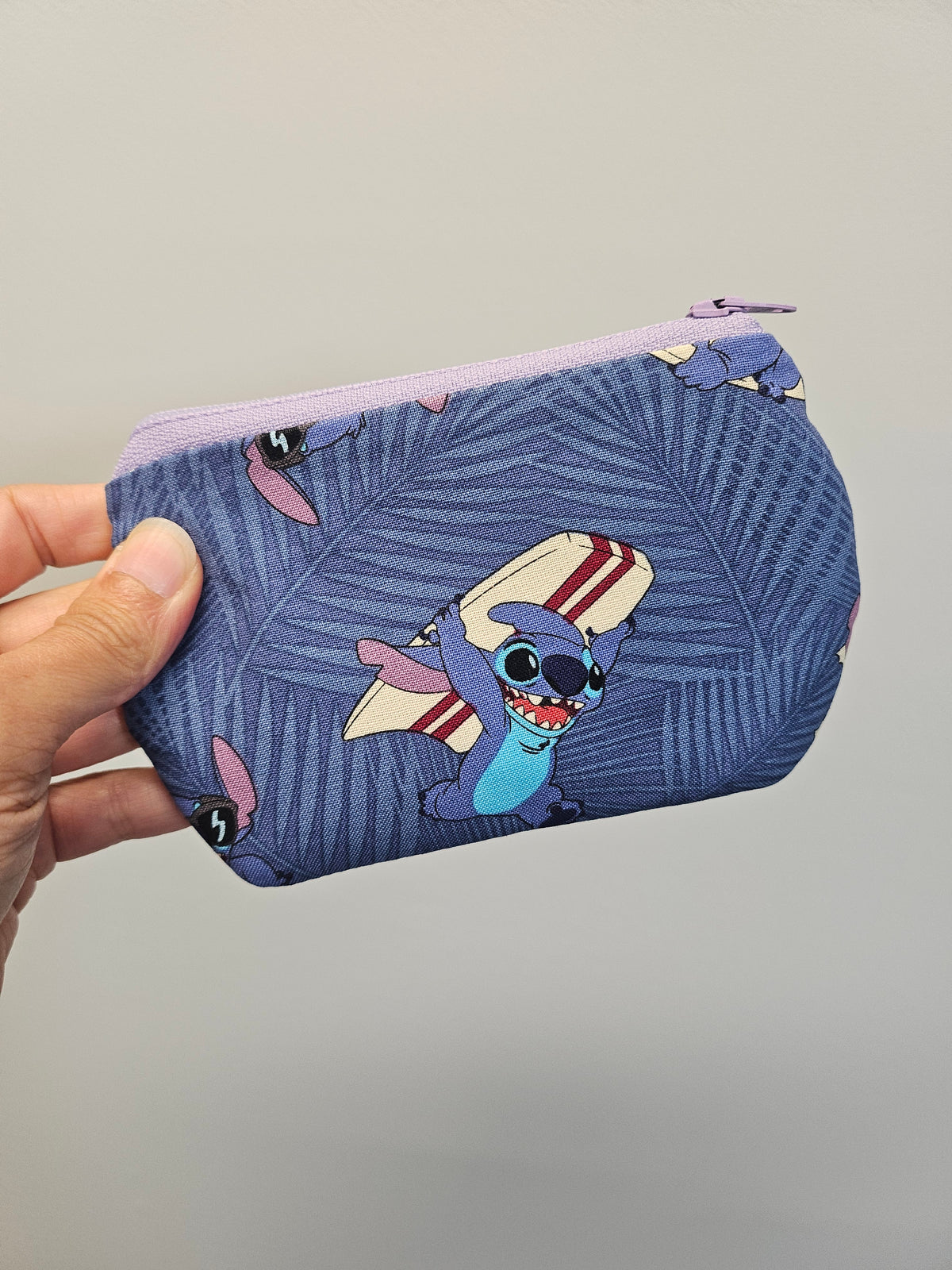 A hand holding a small zippered pouch with a blue tropical leaf pattern and a cartoon character on the front, set against a plain light gray background.