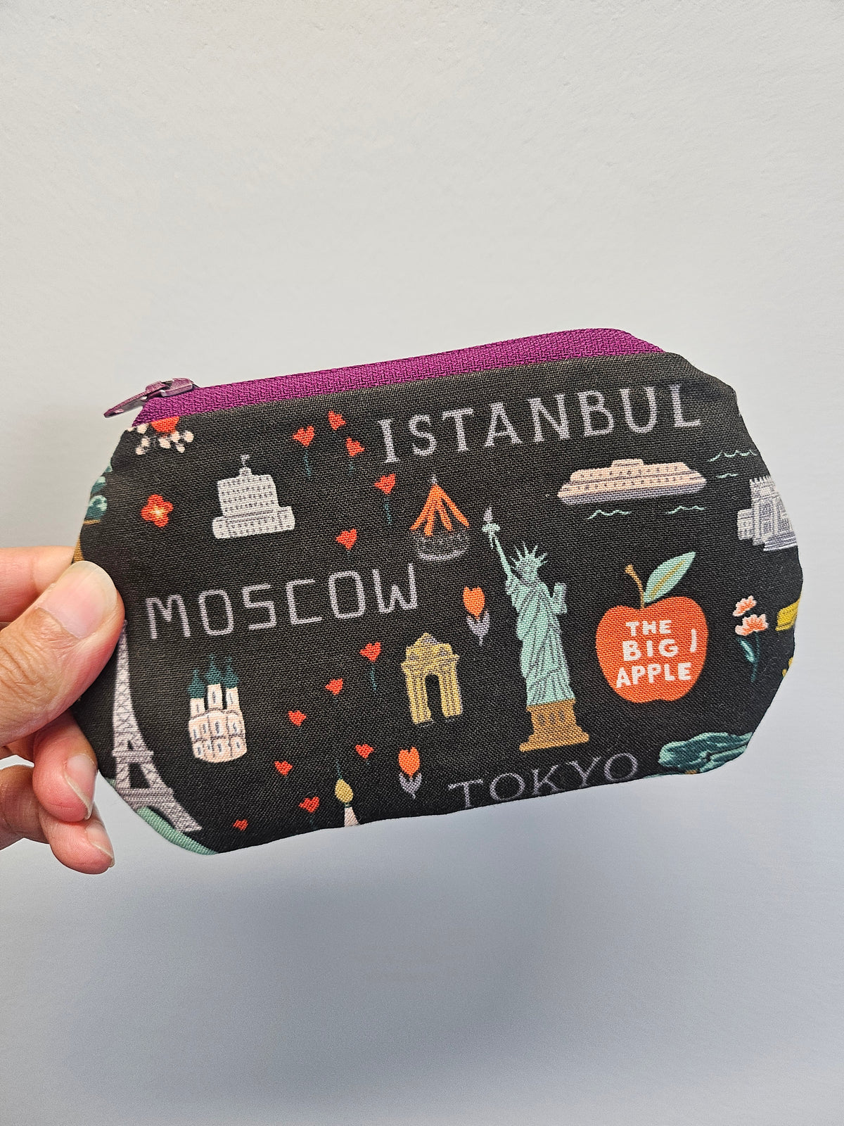 A hand holding a small zippered pouch with a black background printed with travel-themed illustrations and text including Istanbul, Moscow, Tokyo, The Big Apple, and The Statue of Liberty. The pouch has a purple zipper and is photographed against a plain light gray background.