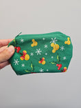 Load image into Gallery viewer, A hand holding a green zippered pouch decorated with a festive holiday print of yellow ducks, red berries, white snowflakes, and green holly leaves against a plain light gray background.
