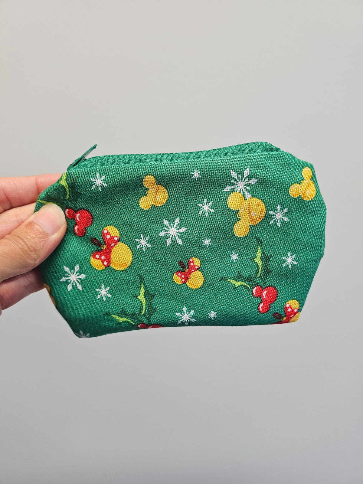 A hand holding a green zippered pouch decorated with a festive holiday print of yellow ducks, red berries, white snowflakes, and green holly leaves against a plain light gray background.