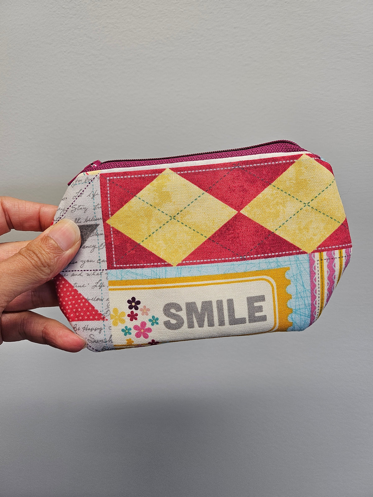 A hand holding a small zippered pouch with a colorful patchwork design, featuring yellow diamond patterns, a red and pink argyle section, and a white panel with the word 'SMILE' and small flowers.