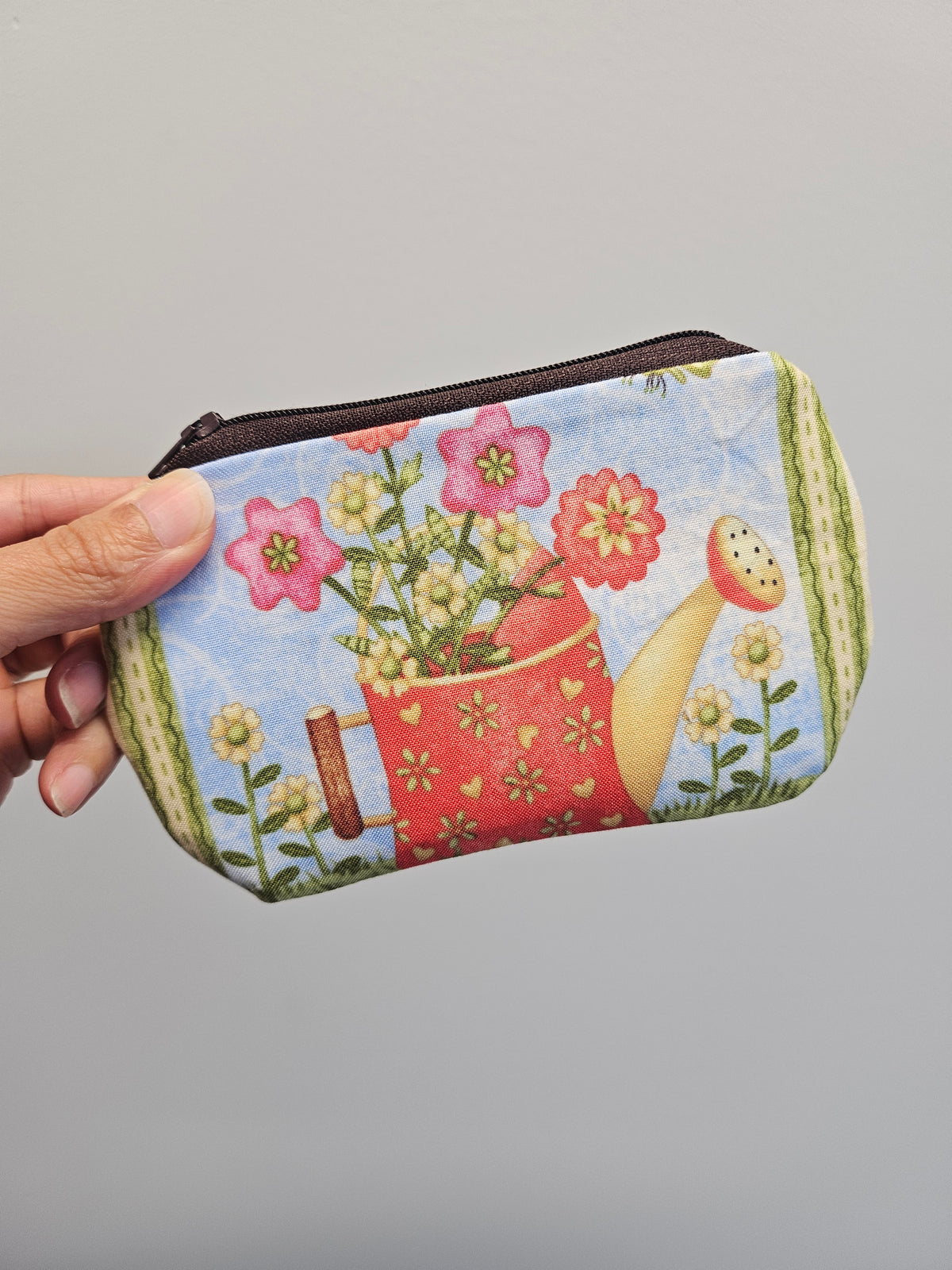 A hand holding a small zippered pouch with a colorful gardening print featuring a red watering can, pink flowers, green leaves, and a light blue background.