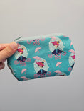 Load image into Gallery viewer, A hand holding a small zippered pouch with a teal background and a repeating pattern of cartoon figures under pink umbrellas.
