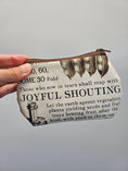 Load image into Gallery viewer, A hand holding a small zippered pouch with a vintage-style printed design featuring text and illustrations, against a plain gray background.
