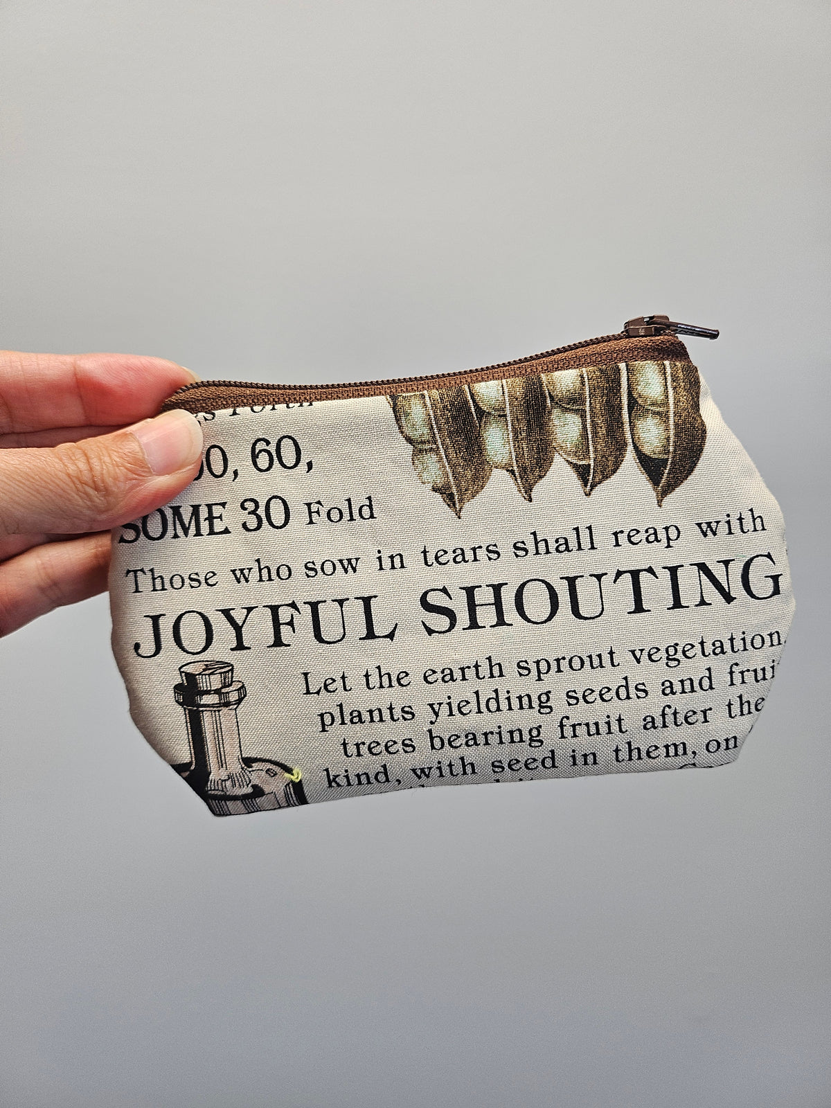 A hand holding a small zippered pouch with a vintage-style printed design featuring text and illustrations, against a plain gray background.