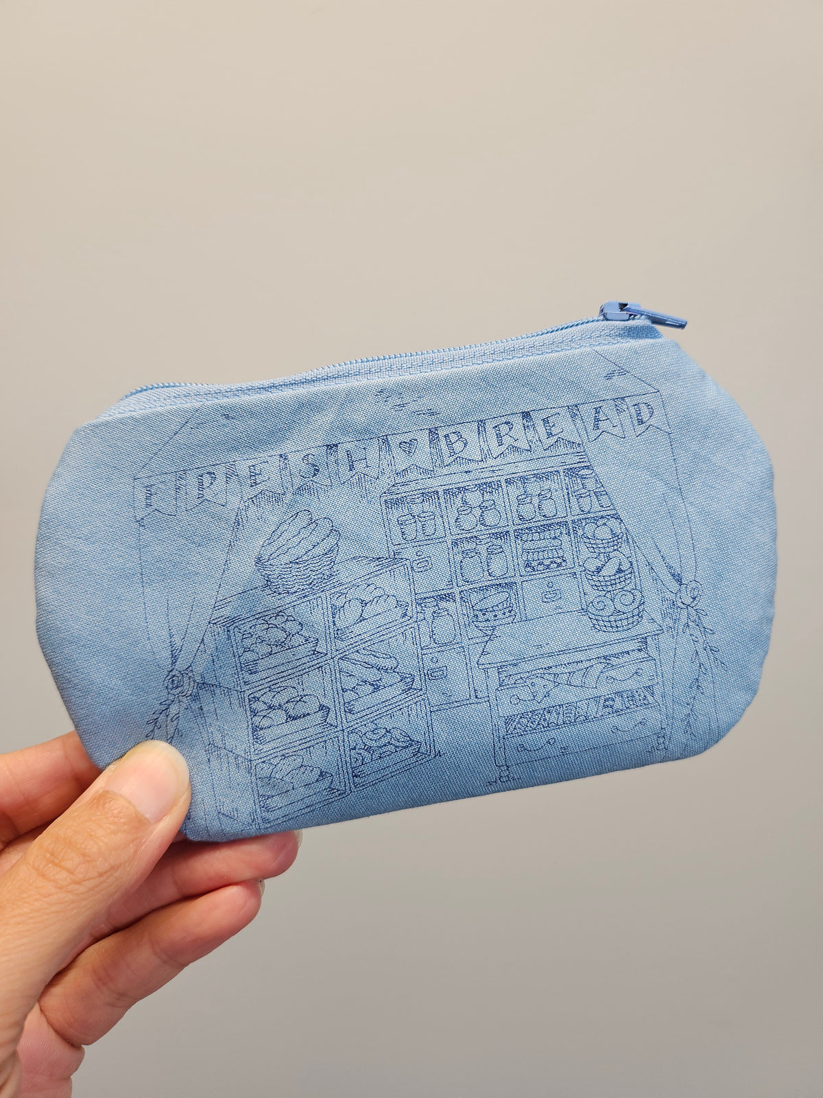 A light blue fabric zippered pouch with a dark blue line-art graphic of a slot machine and the text 'FRESH BREAD' on the front, held by a hand against a plain light gray background.