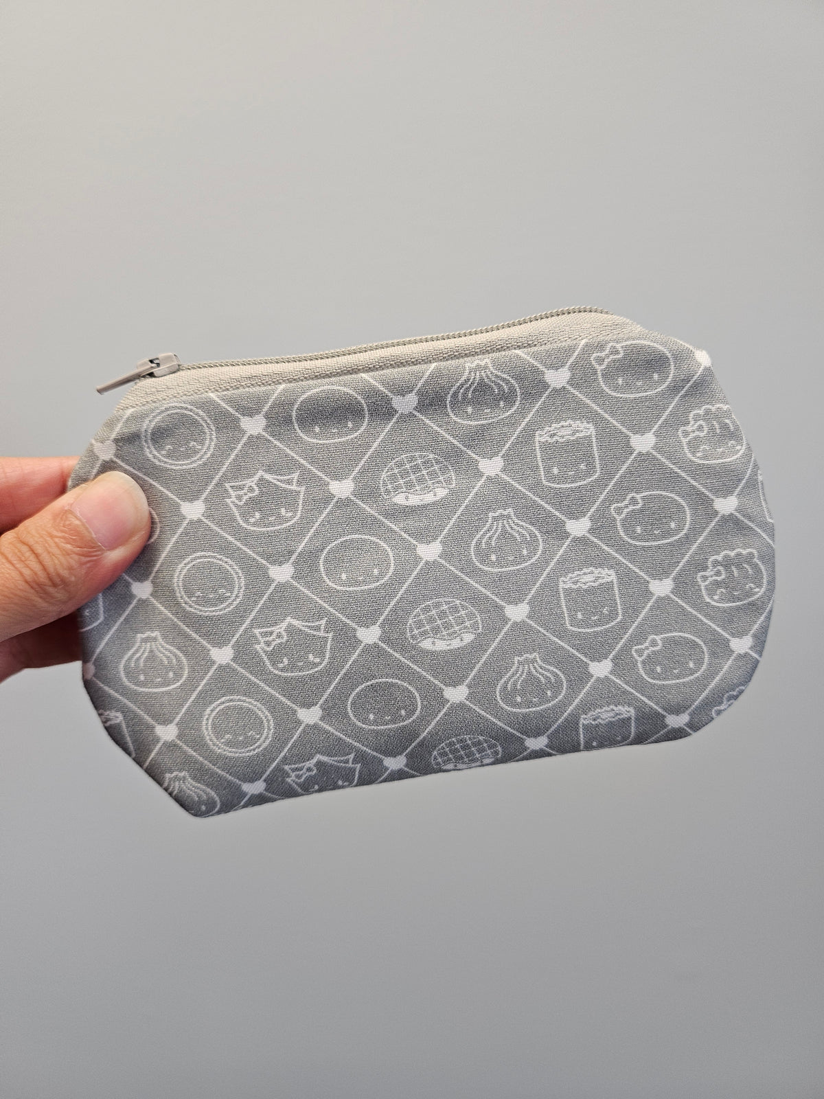 A gray fabric zippered pouch with a white repeating pattern of cute food illustrations, held by a hand against a plain gray background.