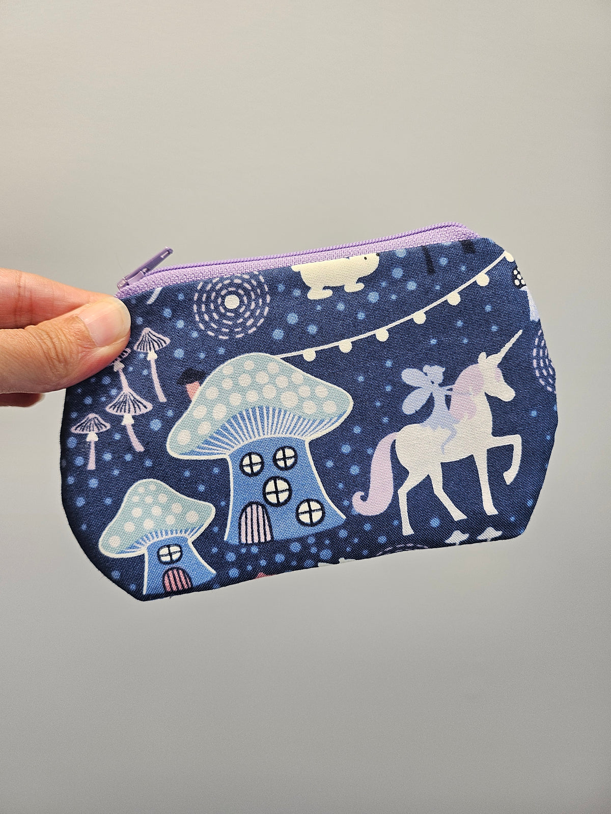 A hand holding a small zippered pouch with a whimsical pattern of blue and white mushrooms, a unicorn, and stars on a dark navy background, set against a plain light gray wall.
