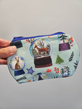 Load image into Gallery viewer, A hand holding a small zippered fabric pouch with a light blue background and a whimsical winter-themed print featuring snow globes, trees, hares, and decorative motifs in multiple colors.
