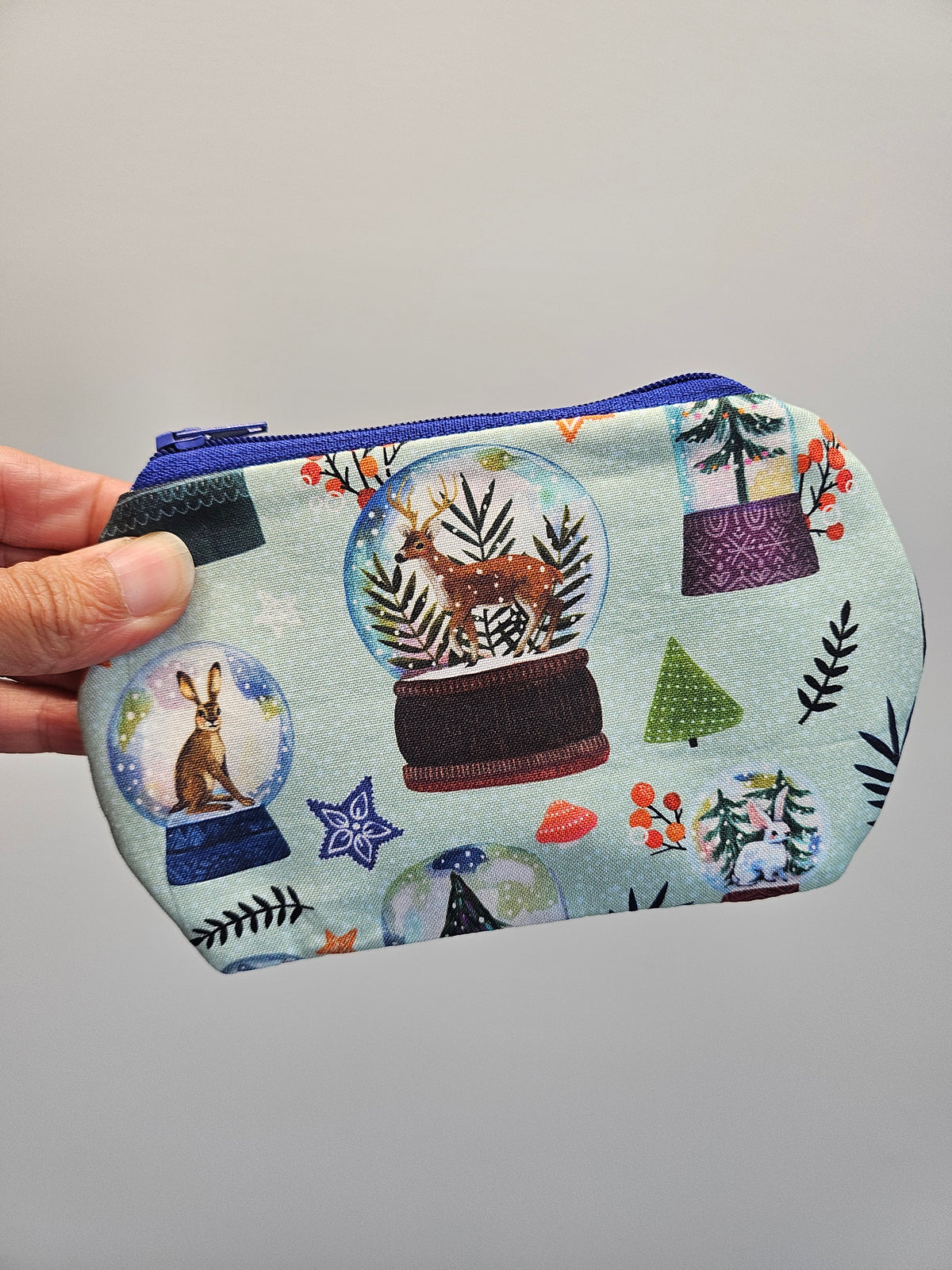 A hand holding a small zippered fabric pouch with a light blue background and a whimsical winter-themed print featuring snow globes, trees, hares, and decorative motifs in multiple colors.