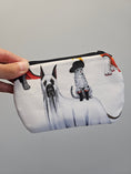 Load image into Gallery viewer, A hand holding a white zippered pouch featuring a printed illustration of a Great Dane in a ghost costume and a small cat wearing a black hat and boots, against a plain gray background.

