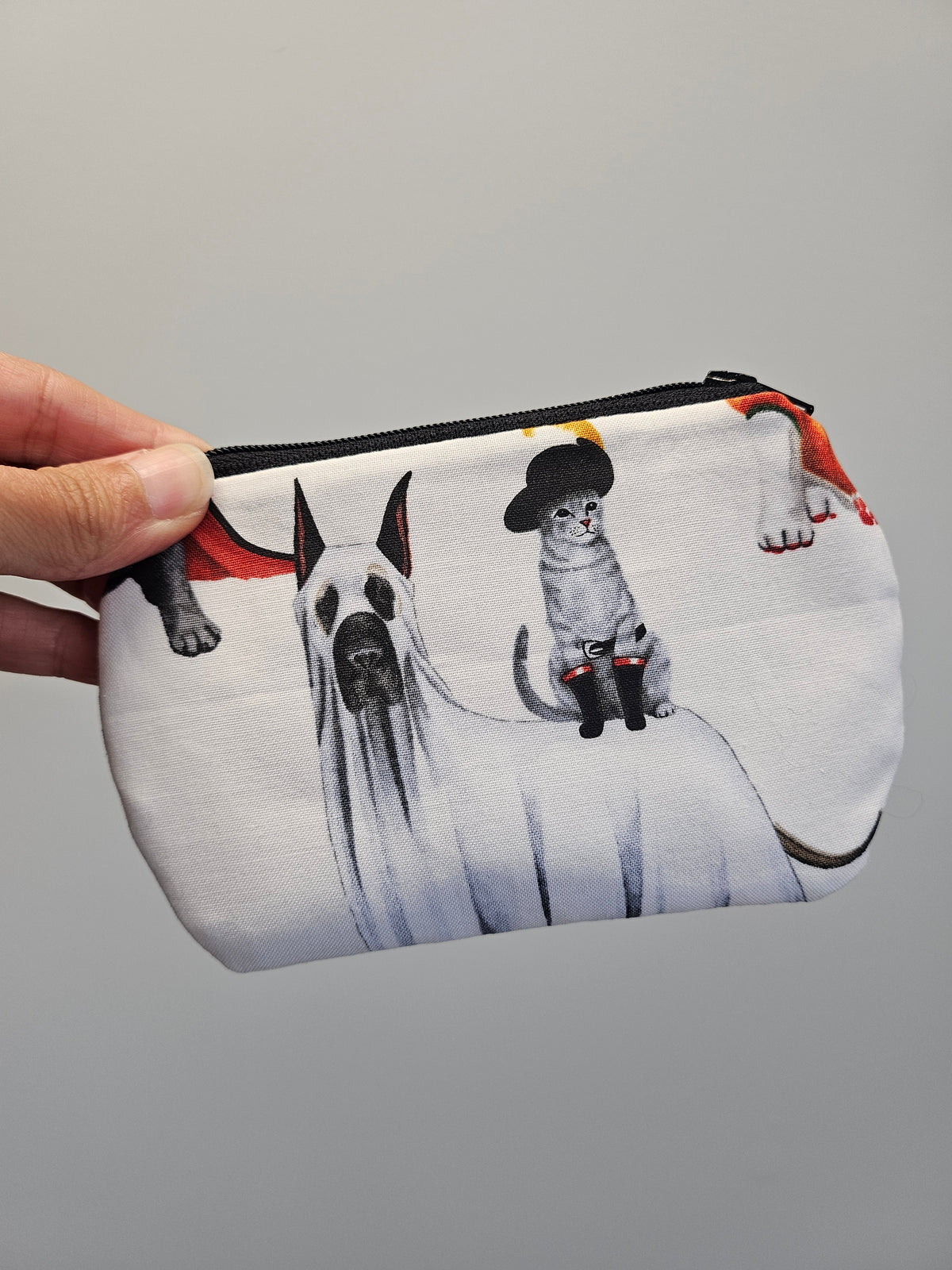 A hand holding a white zippered pouch featuring a printed illustration of a Great Dane in a ghost costume and a small cat wearing a black hat and boots, against a plain gray background.