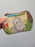 Load image into Gallery viewer, A hand holding a small zippered pouch with a colorful printed design featuring a gray hippopotamus among green plants on a light beige background.
