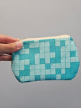 Load image into Gallery viewer, A hand holding a small zippered pouch with a turquoise and light blue crossword puzzle pattern against a plain gray background.
