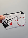 Load image into Gallery viewer, A hand holding a white fabric zippered pouch decorated with medical-themed illustrations of a stethoscope, blood pressure monitor, and heart monitor in black and red.
