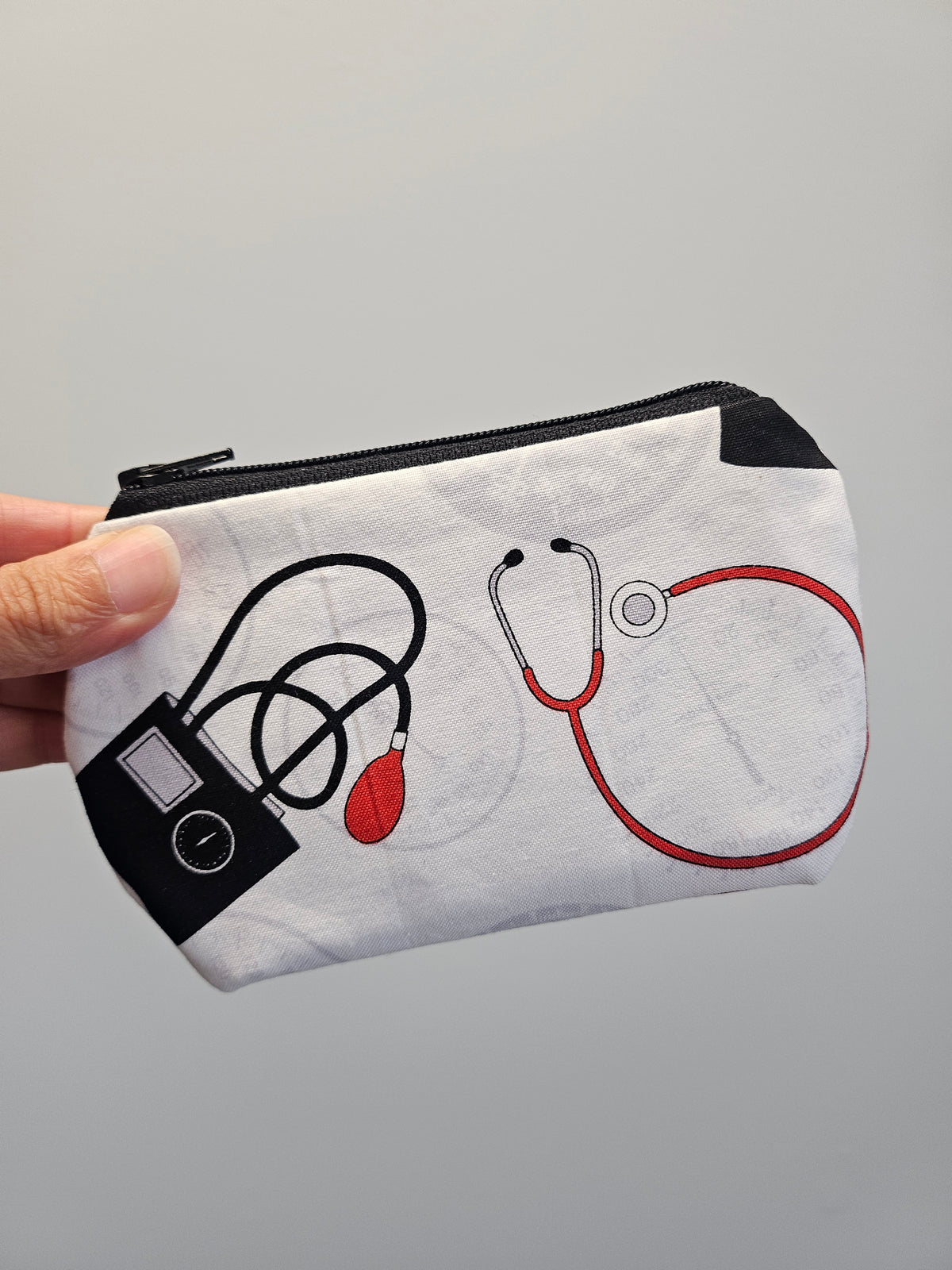 A hand holding a white fabric zippered pouch decorated with medical-themed illustrations of a stethoscope, blood pressure monitor, and heart monitor in black and red.