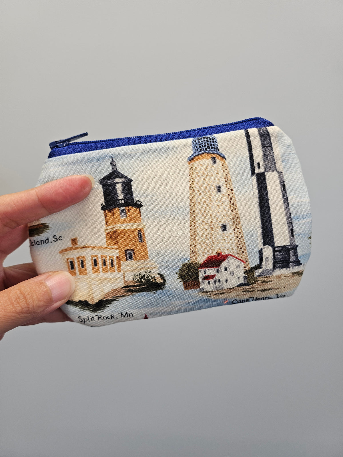 A hand holding a zippered pouch with a printed lighthouse design on a light gray background.