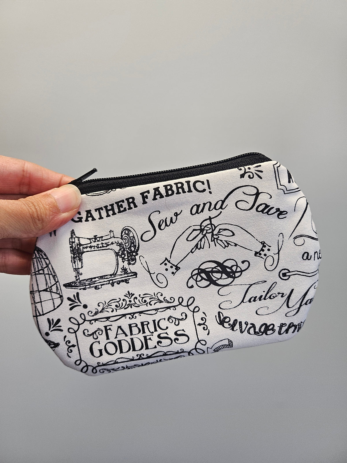 A hand holding a small zippered pouch with a white background and black sewing-themed illustrations and text, including 'GATHER FABRIC!', 'Sew and Save', 'FABRIC GODDESS', and 'Tailor Made'.