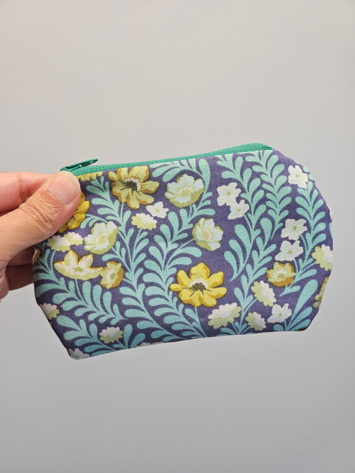 A hand holding a small zippered pouch with a floral pattern. The pouch features a dark purple background with a light blue leafy design, yellow and white flowers, and a mint green zipper.
